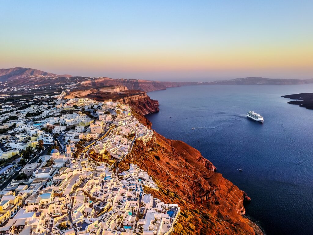 An aerial view of Santorini on the hill top, with white building scattered across the land scape. The ocean could also be seen from this ciew.