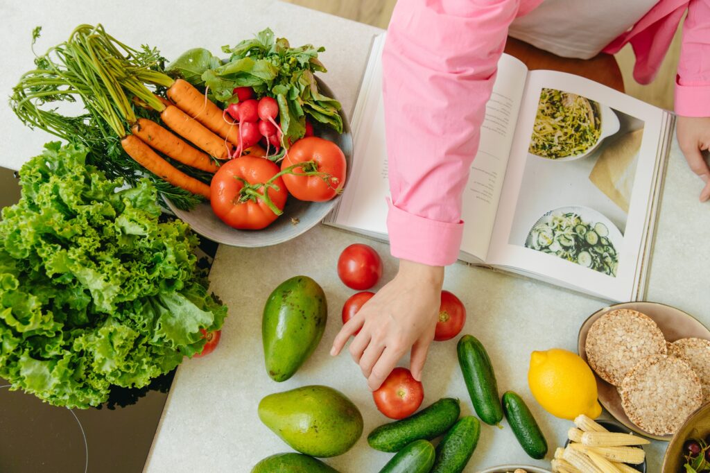 Fruits and vegetables laid out on a table with a cook book as a woman reaches for one of the vegetables.